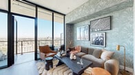 Located in the heart of San Diego at 1401 Union St, this thoughtfully designed high-rise residence offers a compelling blend of contemporary architecture and curated interior design. The living area serves as the centerpiece of the home, featuring dramatic floor-to-ceiling glass walls that frame sweeping panoramic views of the San Diego skyline. Botanical-patterned wallcovering in muted sage tones adds texture and warmth, complemented by a live-edge coffee table, plush area rug, and rich leather accent chairs that create an inviting yet sophisticated atmosphere. Direct access to an outdoor balcony extends the living space further, allowing natural light to flood the interior throughout the day. The kitchen is equipped with stainless steel appliances, warm wood cabinetry, a subway tile backsplash, and a generous quartz-topped island with bar seating, illuminated by modern pendant lighting. A cozy dining nook with a round table and upholstered chairs sits nearby, accented by a striking arched mirror. Wide-plank wood flooring flows consistently through the main living areas, grounding the design with natural warmth. The bathroom features a dual-sink vanity with dark wood cabinetry, white quartz countertops, and illuminated mirrors, conveying a clean, spa-like aesthetic. Every detail of this residence reflects a careful attention to both style and function.