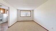 This 2-bedroom, 1-bathroom unit is laid out entirely on a single floor, offering a straightforward and functional floor plan. The space is divided into two bedrooms, a kitchen, a full bathroom, and two hallways that connect the rooms efficiently. The primary bedroom is generously sized relative to the overall footprint, featuring neutral carpet flooring, white walls, and a wood-trimmed window that allows natural light to filter in. A baseboard heating unit runs along the exterior wall, ensuring year-round comfort. The second bedroom is more compact, featuring wood-look flooring, a wood-trimmed window with horizontal blinds, and its own baseboard heater. The full bathroom is finished with white square wall tiles surrounding the tub-shower combination, a wood-framed vanity mirror, chrome fixtures, and a small window that provides natural ventilation and light. The clean white ceiling and walls throughout the unit create a bright and neutral atmosphere. Overall, this unit presents a practical and efficiently organized living space with consistent warm wood accents on window frames and cabinetry, soft neutral tones, and functional baseboard heating in every room.