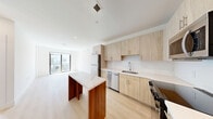 Located at 475 SW 27th Ave in Fort Lauderdale, FL, this modern one-bedroom, two-bathroom unit is part of a contemporary multi-story building with clean architectural lines, balconies, and well-maintained landscaping. Inside, light-toned wood-style flooring runs throughout, complementing the bright white walls and creating a cohesive, modern aesthetic.

The open-concept living and kitchen area serves as the heart of the unit. The kitchen is outfitted with light wood cabinetry, a white subway tile backsplash, stainless steel appliances — including a refrigerator, dishwasher, microwave, and range — and a striking kitchen island featuring a marble-look countertop and warm wood base. Large floor-to-ceiling windows at the end of the living area flood the space with natural light and provide access to a private balcony overlooking the green surroundings.

The bedroom is generously sized, featuring dual windows with black frames that invite abundant natural light and outdoor views, along with a ceiling fan and a double-door closet for ample storage. Two well-appointed bathrooms serve the unit: one with a glass-enclosed walk-in shower and wood-toned vanity, and another with a full soaking tub and tiled surround. A dedicated in-unit laundry room with a full-size washer and dryer completes this efficiently designed and thoughtfully appointed home.