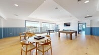 The rec room at 6804 Tides Rd in Rockaway Beach, New York, is a thoughtfully designed amenity space that offers a diverse range of leisure and entertainment areas within a single floor. The interior is unified by light hardwood flooring, crisp white ceilings with recessed lighting, and a coastal-inspired palette accented by powder-blue shiplap wainscoting throughout.

The expansive main lounge features large floor-to-ceiling windows that flood the space with natural light and provide sweeping outdoor views of the surrounding neighborhood. Plush sectional sofas, a round wood coffee table, and geometric pendant light fixtures create a sophisticated yet relaxed gathering environment. An adjacent game area includes a full-size billiards table and shuffleboard table, complemented by casual seating arrangements with woven-back chairs and round tables.

A dedicated screening room offers tiered theater-style seating with reclining leather chairs and a mounted projector, designed for private viewings. A bright, open fitness and yoga studio features a smooth, easy-clean floor and panoramic windows overlooking an outdoor terrace. The catering kitchen is well-equipped with extensive gray cabinetry, white quartz countertops, a herringbone tile backsplash, stainless steel appliances, and a large window overlooking the outdoor deck.