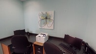 Located at 300 Caughman Farm Ln in Lexington, South Carolina, this single-floor cyber center offers a compact yet well-equipped space designed to meet residents' computing and printing needs. The room features soft teal-painted walls that create a calm, focused atmosphere, complemented by wood-look flooring that adds warmth to the interior.

The space is outfitted with multiple computer workstations, each equipped with a desktop monitor, keyboard, and mouse, positioned along wrap-around dark-surfaced countertops that maximize available desk space. An HP laser printer, set on a dedicated stand with file storage below, is conveniently accessible from multiple stations, making document printing straightforward for users.

A piece of contemporary wall art featuring an abstract floral design adds a decorative touch, softening the utilitarian nature of the room. Posted signage outlines the cyber center's rules and regulations, and a video surveillance notice indicates that the area is monitored for security. The room is accessed through a classic six-panel white door and includes a small waste bin for convenience.

Overall, the cyber center is a practical, neatly organized facility that provides residents with the essential tools for internet access, printing, and general computer use in a tidy and welcoming environment.