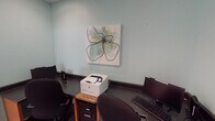 Located at 300 Caughman Farm Ln in Lexington, South Carolina, this single-floor cyber center offers a compact yet well-equipped space designed to meet residents' computing and printing needs. The room features soft teal-painted walls that create a calm, focused atmosphere, complemented by wood-look flooring that adds warmth to the interior.

The space is outfitted with multiple computer workstations, each equipped with a desktop monitor, keyboard, and mouse, positioned along wrap-around dark-surfaced countertops that maximize available desk space. An HP laser printer, set on a dedicated stand with file storage below, is conveniently accessible from multiple stations, making document printing straightforward for users.

A piece of contemporary wall art featuring an abstract floral design adds a decorative touch, softening the utilitarian nature of the room. Posted signage outlines the cyber center's rules and regulations, and a video surveillance notice indicates that the area is monitored for security. The room is accessed through a classic six-panel white door and includes a small waste bin for convenience.

Overall, the cyber center is a practical, neatly organized facility that provides residents with the essential tools for internet access, printing, and general computer use in a tidy and welcoming environment.