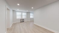 This thoughtfully renovated single-floor space offers a clean, contemporary aesthetic throughout, unified by light wood-tone luxury flooring and soft neutral grey walls that create a calm, inviting atmosphere.

The open living area is generously proportioned, bathed in natural light from multiple windows fitted with white horizontal blinds. Recessed ceiling lighting complements the natural light, and a classic radiator provides efficient heating. The layout provides ample flexibility for a variety of furniture arrangements.

The kitchen is efficiently designed with a functional galley-style layout. It features two-tone cabinetry—crisp white upper cabinets paired with dark lower cabinets—topped with light quartz countertops. Stainless steel appliances, including a full-size refrigerator, an electric range with overhead microwave, and a stainless sink, complete this well-appointed space. A tile backsplash with a wood-grain pattern adds a warm, modern touch.

The bedroom offers a comfortable footprint with a built-in closet, recessed lighting, and a window allowing in natural light. Clean white Shaker-style doors and trim throughout maintain the unit's cohesive, modern design language.

The full bathroom is elegantly finished with large-format light tile flooring, white subway tile walls accented by a decorative mosaic tile border, a glass-enclosed walk-in shower with a built-in bench, and a dark wood vanity. A small window provides natural ventilation. An additional open room offers versatile space suitable for a home office or flex room.