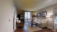 This single-floor residence offers a thoughtfully designed living space that blends comfort and contemporary style. The living room serves as the heart of the home, showcasing rich hardwood flooring that anchors the space with warmth and elegance. Neutral taupe wall tones create a versatile backdrop, complemented by a tufted cream sofa, a woven accent chair, and a dark wood coffee table that together form a cohesive and inviting seating arrangement. A natural fiber area rug defines the conversation area, adding texture and depth to the room. A dark wood display cabinet provides both storage and visual interest along one wall, while framed artwork adds a curated, polished touch to the décor. The living room flows seamlessly toward a sliding glass door dressed in floor-length draping curtains, opening onto a private balcony that captures abundant natural light and offers views of the surrounding cityscape. The overall ambiance is calm and refined, with a harmonious balance of natural materials and neutral tones that create a welcoming, move-in-ready atmosphere throughout the space.