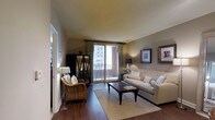 This single-floor residence offers a thoughtfully designed living space that blends comfort and contemporary style. The living room serves as the heart of the home, showcasing rich hardwood flooring that anchors the space with warmth and elegance. Neutral taupe wall tones create a versatile backdrop, complemented by a tufted cream sofa, a woven accent chair, and a dark wood coffee table that together form a cohesive and inviting seating arrangement. A natural fiber area rug defines the conversation area, adding texture and depth to the room. A dark wood display cabinet provides both storage and visual interest along one wall, while framed artwork adds a curated, polished touch to the décor. The living room flows seamlessly toward a sliding glass door dressed in floor-length draping curtains, opening onto a private balcony that captures abundant natural light and offers views of the surrounding cityscape. The overall ambiance is calm and refined, with a harmonious balance of natural materials and neutral tones that create a welcoming, move-in-ready atmosphere throughout the space.