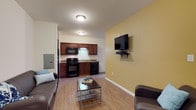 This single-floor unit offers an efficient and functional layout with an open-concept design that seamlessly connects the living area and kitchen. The living space features warm hardwood-style flooring that extends throughout, providing a cohesive and clean aesthetic. A bold yellow accent wall adds a touch of warmth and color to the room, complementing the neutral white walls. The living area is well-suited for relaxation, with ample space for seating and a wall-mounted flat-screen TV with a dedicated media shelf, keeping the setup sleek and organized. The kitchen is positioned directly adjacent to the living area and is equipped with rich dark wood cabinetry, black appliances including a range, built-in microwave, and dishwasher, along with a functional sink and countertop workspace. Flush-mount ceiling lighting provides consistent illumination throughout the space. An electrical panel is conveniently accessible along one wall. The overall ambiance is practical and comfortable, with a straightforward design that maximizes the use of available space, making it a well-appointed and move-in ready unit.