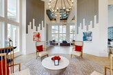 Located at 13030 Perry Rd in Houston, TX, this thoughtfully designed community clubhouse offers a sophisticated and welcoming environment across a single, generously sized floor. The grand entrance lobby immediately impresses with soaring double-height ceilings, dramatic vertical shiplap wall paneling in gray and white, and a striking cityscape-inspired wall motif. A large-scale brass chandelier anchors the space, casting warm light over gold-framed wingback chairs, a marble-topped round coffee table, and curated abstract artwork. Expansive floor-to-ceiling windows flood the interior with natural light and offer views of an outdoor pool and palm-lined grounds. The adjacent lounge features a bold teal accent wall with a mounted TV, plush seating, and continued views of the pool area, creating a seamless indoor-outdoor connection. A stylish café bar showcases a stunning hexagonal teal tile backsplash, marble countertops, and pendant lighting above bar-height seating. A cozy dining nook with channel-tufted banquette seating against a quilted teal accent wall provides an intimate gathering spot. Light hardwood flooring flows throughout, unifying each area. A bright, open office space with high ceilings, large windows, and built-in shelving completes this well-appointed clubhouse.