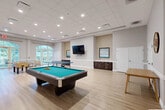 Located at 2855 W Commercial Blvd in Fort Lauderdale, Florida, this generously sized single-floor game and recreation room offers a well-appointed space designed for leisure and social activity. The open layout accommodates a full-size pool table with cue rack, a ping-pong table, and a foosball table, providing a variety of recreational options within a single, cohesive environment.

The room features light hardwood-style flooring throughout, contributing to a bright and clean aesthetic. A drop tile ceiling with recessed lighting ensures even, consistent illumination across the space. On the main feature wall, a large wall-mounted flat-screen television is flanked by textured accent panels and framed coastal artwork, with dark wood built-in cabinetry below providing additional storage. White chair rail molding lines the perimeter walls, adding a classic architectural detail.

A seating area near the entrance features large arched French doors and arched transom windows that flood the space with natural light and offer views of the surrounding tropical greenery. Additional seating is available throughout the room, including a wooden bench and small accent tables. A whiteboard panel and wall clock are also present, enhancing the room's functionality. The overall ambiance is clean, modern, and inviting — well-suited for community recreation.