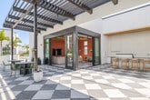 Located at 3988 E Hardy St in Inglewood, California, this single-level amenity space combines a thoughtfully designed interior lounge with a generous outdoor courtyard, offering a well-rounded environment for gathering and relaxation. The outdoor areas are defined by a striking checkerboard-patterned tile floor that extends beneath a series of dark-framed pergolas, casting dynamic shadow patterns across the space throughout the day. Multiple seating arrangements are distributed across the courtyard, including modular sofas, fire pit tables with wooden surround seating, and café-style dining sets. String lights are strung overhead, adding ambiance for evening use, while mature palm trees and curated landscaping with raised planters introduce lush greenery throughout. A separate lounge zone features low-profile seating on a grass lawn, adding a more casual outdoor retreat. The interior lounge is characterized by warm terracotta-toned shiplap walls, exposed wood ceiling beams, and a bold checkerboard floor. Abundant natural light enters through multiple clerestory and standard windows, illuminating a mix of swivel lounge chairs, sofas, and bar-height seating. Large glass sliding doors seamlessly connect the interior lounge to the outdoor courtyard, creating a cohesive and inviting amenity experience.