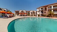 Located at 4722 E Bell Rd in Phoenix, Arizona, this outdoor amenity space showcases a resort-style pool environment designed for relaxation and recreation under the Arizona sun. The centerpiece is a generously sized freeform swimming pool with clear, blue-green water, complemented by a broad surrounding sun deck finished in textured aggregate concrete. The pool area is well-appointed with rows of blue lounge chairs shaded by vibrant orange umbrellas, creating a striking and inviting color palette against the bright desert sky. Tall, mature palm trees are positioned throughout the space, providing natural shade and reinforcing the resort ambiance. The surrounding buildings reflect a Spanish Colonial Revival architectural style, characterized by white stucco exteriors, terracotta tile rooflines, arched balconies, and decorative ironwork. A covered structure with a clay tile roof serves as a transition point between the pool deck and an interior facility, featuring large multi-pane windows that look out onto the pool. Additional recreational items, such as pool floats, are visible near building entrances. The overall atmosphere is bright, open, and ideally suited to the warm Phoenix climate, offering a well-designed outdoor retreat with cohesive architectural and landscaping elements throughout.
