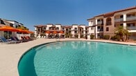 Located at 4722 E Bell Rd in Phoenix, Arizona, this outdoor amenity space showcases a resort-style pool environment designed for relaxation and recreation under the Arizona sun. The centerpiece is a generously sized freeform swimming pool with clear, blue-green water, complemented by a broad surrounding sun deck finished in textured aggregate concrete. The pool area is well-appointed with rows of blue lounge chairs shaded by vibrant orange umbrellas, creating a striking and inviting color palette against the bright desert sky. Tall, mature palm trees are positioned throughout the space, providing natural shade and reinforcing the resort ambiance. The surrounding buildings reflect a Spanish Colonial Revival architectural style, characterized by white stucco exteriors, terracotta tile rooflines, arched balconies, and decorative ironwork. A covered structure with a clay tile roof serves as a transition point between the pool deck and an interior facility, featuring large multi-pane windows that look out onto the pool. Additional recreational items, such as pool floats, are visible near building entrances. The overall atmosphere is bright, open, and ideally suited to the warm Phoenix climate, offering a well-designed outdoor retreat with cohesive architectural and landscaping elements throughout.