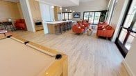 This single-floor amenity space is designed for both relaxation and entertainment, combining multiple functional areas within one open, airy layout. Light hardwood-style flooring runs throughout, lending the space a clean and contemporary feel that ties each zone together seamlessly.

At the center of the space sits a full kitchen equipped with stainless steel appliances, including a built-in wall oven and a spacious refrigerator. A large kitchen island with a white countertop is lined with sleek, modern bar stools, creating an inviting spot for casual dining or conversation. Pendant lighting hangs above the island, adding a warm, focused glow to the area. Warm wood cabinetry complements the light tones of the flooring throughout.

Adjacent to the kitchen, a lounge area is furnished with bold orange upholstered chairs arranged in a social configuration, anchored by a wall-mounted flat-screen television. Large sliding glass doors and generous windows flood this corner with natural light and provide views of the outdoor greenery beyond.

In the foreground, a billiards table adds a recreational dimension to the space. Together, these elements create a well-rounded, welcoming communal environment suited for a variety of gatherings.