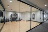 Located at 3 Hawkins Ave in Ronkonkoma, NY, this upscale amenity lounge offers a thoughtfully designed, open-concept floor spanning a generous single level. The space is defined by its contemporary aesthetic, blending light hardwood flooring, crisp white ceilings with linear beam detailing, and striking black steel-framed glass partitions that divide the floor into distinct yet visually connected zones.

The main lounge area features multiple seating arrangements to accommodate a variety of uses, including a bold mustard-yellow sectional sofa positioned beneath a multi-arm pendant light and flanked by large windows that fill the space with abundant natural light and outdoor views. Additional seating vignettes offer intimate conversation areas with stylish accent chairs and round tables throughout.

A sleek bar runs along one side of the space, lined with dark green upholstered barstools and illuminated by globe pendant fixtures, complemented by a bold geometric tile floor detail. A private conference room, enclosed by glass partitions, serves as a standout feature — anchored by a dark round table, upholstered chairs, and a dramatic sculptural gold chandelier that casts intricate shadow patterns across the grey walls. A separate lounge alcove with a channeled leather sofa and curated artwork adds a refined, gallery-like touch to the overall ambiance.