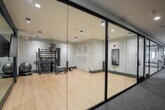 Located at 3 Hawkins Ave in Ronkonkoma, NY, this upscale amenity lounge offers a thoughtfully designed, open-concept floor spanning a generous single level. The space is defined by its contemporary aesthetic, blending light hardwood flooring, crisp white ceilings with linear beam detailing, and striking black steel-framed glass partitions that divide the floor into distinct yet visually connected zones.

The main lounge area features multiple seating arrangements to accommodate a variety of uses, including a bold mustard-yellow sectional sofa positioned beneath a multi-arm pendant light and flanked by large windows that fill the space with abundant natural light and outdoor views. Additional seating vignettes offer intimate conversation areas with stylish accent chairs and round tables throughout.

A sleek bar runs along one side of the space, lined with dark green upholstered barstools and illuminated by globe pendant fixtures, complemented by a bold geometric tile floor detail. A private conference room, enclosed by glass partitions, serves as a standout feature — anchored by a dark round table, upholstered chairs, and a dramatic sculptural gold chandelier that casts intricate shadow patterns across the grey walls. A separate lounge alcove with a channeled leather sofa and curated artwork adds a refined, gallery-like touch to the overall ambiance.