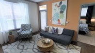 This single-floor space offers a thoughtfully designed interior that balances comfort and contemporary style. The living area serves as the heart of the space, featuring a warm terracotta-orange accent wall that creates an inviting, energetic focal point. A modern charcoal futon sofa, layered with an assortment of patterned and textured throw pillows, anchors the seating area alongside a bold chevron-patterned accent chair that adds visual interest and personality. A rustic round wooden coffee table with intricate detailing grounds the space and complements the geometric-patterned area rug beneath. A large, vibrant abstract canvas artwork adorns the orange accent wall, introducing splashes of teal, green, and orange that tie the room's color palette together cohesively. Natural light flows generously through multiple windows dressed with white sheer curtains and horizontal blinds, creating a bright and airy atmosphere. A glass sliding or hinged door provides access to an outdoor area, further enhancing the connection between interior and exterior. The dark hardwood flooring adds warmth and contrast to the lighter furnishings, while neutral gray walls on the remaining surfaces keep the overall aesthetic balanced and modern. The space presents a functional yet stylish environment with careful attention to decor detail.