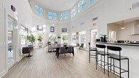 This single-story clubhouse in Columbia, Maryland, is designed to serve as a welcoming community gathering space, combining contemporary design with functional amenities. The architecture immediately impresses with dramatically soaring ceilings in the main gathering area, encircled by a band of large clerestory windows that flood the interior with natural light throughout the day. Crisp white walls and light wood-look flooring extend throughout, creating a clean, airy aesthetic that feels both modern and inviting.

The expansive main lounge area is thoughtfully arranged with multiple seating configurations, including a large dining-style table with upholstered chairs suited for group gatherings, as well as a casual lounge corner featuring accent chairs, a bench, and potted fiddle-leaf fig trees that add organic warmth to the space. Large exterior-facing windows and glass-paneled doors provide views of the surrounding outdoor areas, reinforcing a strong connection between interior and exterior.

A well-appointed kitchen sits adjacent to the main gathering area, outfitted with stainless steel appliances including a range, microwave, and dishwasher, complemented by dark granite countertops and white cabinetry. A peninsula with bar-height seating extends into the social space, making it ideal for casual dining or entertaining. The overall ambiance is bright, polished, and community-focused.
