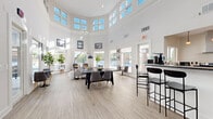 This single-story clubhouse in Columbia, Maryland, is designed to serve as a welcoming community gathering space, combining contemporary design with functional amenities. The architecture immediately impresses with dramatically soaring ceilings in the main gathering area, encircled by a band of large clerestory windows that flood the interior with natural light throughout the day. Crisp white walls and light wood-look flooring extend throughout, creating a clean, airy aesthetic that feels both modern and inviting.

The expansive main lounge area is thoughtfully arranged with multiple seating configurations, including a large dining-style table with upholstered chairs suited for group gatherings, as well as a casual lounge corner featuring accent chairs, a bench, and potted fiddle-leaf fig trees that add organic warmth to the space. Large exterior-facing windows and glass-paneled doors provide views of the surrounding outdoor areas, reinforcing a strong connection between interior and exterior.

A well-appointed kitchen sits adjacent to the main gathering area, outfitted with stainless steel appliances including a range, microwave, and dishwasher, complemented by dark granite countertops and white cabinetry. A peninsula with bar-height seating extends into the social space, making it ideal for casual dining or entertaining. The overall ambiance is bright, polished, and community-focused.