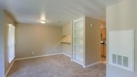 This single-floor space offers a clean, functional layout with a neutral color palette throughout. The main living area features wall-to-wall carpeting and warm beige walls, creating a comfortable and inviting atmosphere. A standout feature of the space is the built-in open bookshelf unit, which provides ample display and storage space without occupying additional floor area. Adjacent to the shelving, a built-in desk nook with overhead cabinetry offers a dedicated workspace seamlessly integrated into the living area — a practical detail that adds versatility to the room. Flush-mount ceiling lighting provides consistent illumination across the space. Transitioning from the living area, the kitchen is visible and features warm wood-toned cabinetry, a countertop with appliances, and hard flooring — contrasting the carpeted areas and providing a durable, easy-to-maintain surface suited for a cooking environment. White trim and baseboards throughout the space provide crisp, clean definition between the walls and floors. Overall, the space presents a straightforward and efficient design, well-suited for everyday living with thoughtful built-in features that maximize functionality.