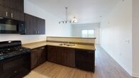This well-designed single-floor unit offers a clean, contemporary aesthetic with a cohesive palette of warm wood-look flooring and crisp white walls carried throughout the entire space. The open-concept kitchen is a standout feature, equipped with dark espresso-toned cabinetry that extends to the ceiling for ample storage, a full suite of black appliances including a gas range, over-the-range microwave, refrigerator, and dishwasher, and a double-basin sink set into a neutral-toned countertop. A peninsula-style breakfast bar separates the kitchen from the living area, which is illuminated by a stylish track and pendant lighting combination. The living area flows naturally toward a sliding glass door that opens to a balcony with open outdoor views. The bedrooms are bright and airy, each benefiting from generous windows that invite abundant natural light and exterior views. The two bathrooms are finished consistently with the rest of the unit, featuring dark wood-toned vanities with neutral countertops, wood-look flooring, and full tub-shower combinations with clean white surrounds. Overall, the unit presents a modern, move-in-ready environment with thoughtful design details and a bright, open ambiance throughout.