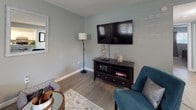 This single-floor residence offers a thoughtfully designed layout that balances comfort and functionality. The kitchen is a standout feature, showcasing white shaker cabinetry, granite countertops, a mosaic tile backsplash in blue and grey tones, stainless steel appliances including a dishwasher and refrigerator, and open shelving. A pass-through window connects the kitchen directly to the living area, promoting an open, social atmosphere. The living room is a comfortable, light-filled space with wood-look flooring, soft grey walls, and a wall-mounted flat-screen TV. The adjacent dining area benefits from natural light through large windows and features chair rail molding, adding a classic architectural detail. Both bedrooms are carpeted and dressed in calming sage-green tones. The primary bedroom offers sliding glass door access with outdoor views of lush greenery, while the second bedroom features a window with natural light. The bathroom is well-equipped with a granite vanity top, white cabinetry, wood-look flooring, and a full tub-shower combination. Throughout the residence, a consistent palette of soft greys, greens, and warm neutrals creates a cohesive and inviting ambiance.