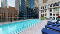 Located atop 29 S La Salle Street in the heart of Chicago's Loop, this rooftop pool deck offers a distinctive urban retreat surrounded by the city's iconic skyline. The centerpiece is a generously sized lap pool with crystal-clear blue water, framed by sleek stainless steel railings and accessible via multiple entry points. The pool is bordered by a spacious tiled sun deck outfitted with navy blue chaise lounges, providing ample space for relaxation while taking in panoramic views of neighboring glass skyscrapers and historic Chicago architecture.

A shaded pergola area with modern black framing provides a comfortable lounge zone, furnished with curved light-gray sectional seating, teal accent poufs, and additional chaise lounges beneath pendant lighting. Lush greenery is thoughtfully integrated throughout the space, including a dramatic living plant wall featuring a diverse array of ferns, succulents, and tropical foliage that frames a dramatic arched entryway. Potted plants and vertical garden elements add vibrant contrast to the clean, neutral palette of the tile deck.

The overall ambiance balances sophisticated urban design with resort-style comfort, offering an inviting outdoor escape in the middle of one of Chicago's most dynamic downtown corridors.