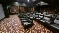 This purpose-built home theater is designed to deliver a true cinematic experience in a private, controlled environment. The room is finished in deep, dark tones throughout — from the dark walls and ceiling to the rich, patterned carpet — creating an immersive atmosphere that minimizes light distraction and enhances focus on the screen. Multiple rows of plush, full-sized black leather recliner seats are arranged in a tiered layout, ensuring unobstructed sightlines from every position in the room. Each recliner features integrated cup holders, combining comfort with practicality for extended viewing sessions. The walls are lined with decorative acoustic panels framed in warm tones, which serve both an aesthetic and functional purpose by managing sound reflection and absorption for optimized audio quality. A professional ceiling-mounted projector is positioned at the rear of the room, aimed toward the front screen. Recessed ceiling lighting provides subtle ambient illumination when needed. The boldly patterned carpet in deep red and gold tones adds a classic, theater-inspired elegance to the space, tying together the overall design aesthetic. This theater is a standout space built for serious film enthusiasts.