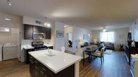 This single-floor residence is designed around an open-concept floor plan that unifies the kitchen, dining, and living spaces into one cohesive, flowing environment. The kitchen is outfitted with dark espresso cabinetry, stainless steel appliances including a gas range and built-in microwave, and a large island topped with a bright white quartz countertop that serves as both a prep area and casual gathering spot. Pendant lighting accents the kitchen, adding warmth and visual interest overhead.

The dining area sits adjacent to the kitchen, furnished with a dark rectangular table and upholstered chairs, creating a defined yet open space for meals. Hardwood-style flooring extends throughout, lending consistency and a warm aesthetic across the common areas.

The living room occupies the far end of the open layout, benefiting from natural light streaming through large windows dressed with sheer curtains. A ceiling fan provides additional comfort. Neutral tones dominate the walls and furnishings, creating a calm, welcoming ambiance. A hallway visible from the main space leads to additional rooms, suggesting a well-organized and functional floor plan throughout.