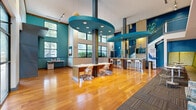 This striking single-floor community space makes an immediate impression with its soaring double-height ceilings and an abundance of natural light streaming through expansive floor-to-ceiling windows. The design is anchored by a bold, contemporary color palette — deep teal walls, chartreuse accents, and warm hardwood flooring — creating a lively yet cohesive atmosphere throughout.

The central area features polished wood-and-stone bar-height counters with white ergonomic seating, ideal for casual work or socializing. Distinctive circular teal ceiling fixtures with integrated lighting serve as dramatic focal points overhead. A wet bar and beverage station add a hospitality-forward touch to the space.

Lounge seating areas are thoughtfully arranged throughout, offering plush sofas, modern armchairs, and intimate booth-style nooks upholstered in metallic gold fabric. A media wall clad in bold teal patterned tile houses a mounted flat-screen television, creating a cozy entertainment corner. Curated abstract artwork adorns the vivid blue accent walls, adding a gallery-like quality.

Large windows frame urban views, flooding the space with daylight and connecting the interior to the surrounding cityscape. The combination of warm hardwood, patterned carpet tiles, tile-clad columns, and pendant lighting creates a dynamic, welcoming environment suited for both relaxed lounging and social gatherings.