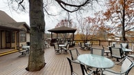 The Clubhouse Deck is a generously sized outdoor gathering space designed to accommodate multiple groups simultaneously. The deck is constructed with wood planking and is enclosed by a dark-railed perimeter fence, providing both safety and a defined boundary while preserving open sightlines to the surrounding tree-lined landscape. Mature trees frame the space throughout, offering natural character and partial shade.

The deck is organized across two levels connected by a short staircase. The lower, primary level hosts numerous round glass-top tables paired with swivel patio chairs, distributed across the open expanse to allow flexible seating arrangements. A gas grill is positioned along one side, making the space well-suited for outdoor gatherings and events. The upper level features a prominent wood gazebo with a pitched roof, providing a sheltered seating area with elevated views of the natural surroundings.

Adjacent to the clubhouse building, a covered porch with decorative columns and French doors provides a seamless transition between the interior and the deck. The overall ambiance is relaxed and communal, enhanced by the natural wooded backdrop and open-air setting.