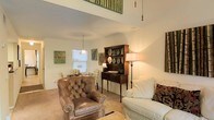 This two-floor space offers a thoughtfully designed interior that combines classic elegance with warm, homey comfort. The living area immediately draws the eye upward with its impressive double-height ceiling, creating an expansive, airy atmosphere that feels both grand and welcoming. A mezzanine-style upper level overlooks the main living space below, adding architectural interest and a sense of vertical depth.

The main living area is appointed with a rich mix of traditional furnishings, including a tufted leather armchair, a plush white sofa, and a stately dark wood hutch displaying decorative dishware — all contributing to a refined yet relaxed aesthetic. Warm-toned walls and light carpeting create a soft, neutral backdrop that enhances the natural flow of the space. Pendant lighting and freestanding floor lamps provide layered illumination, casting a warm glow throughout the room.

Botanical and nature-inspired artwork adorns the walls, reinforcing a serene, organic ambiance. A window in the living area allows natural light to filter in, brightening the space. The overall design reflects a timeless, traditional sensibility, with carefully curated décor details that lend the space a lived-in warmth and a sense of enduring character across both levels.