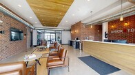 Located at 731 NE Venture Dr in Waukee, Iowa, this amenity center at Brick Towne offers a thoughtfully designed single-floor layout spanning a generous footprint. The space blends industrial and modern aesthetics, highlighted by exposed brick accent walls, polished concrete floors, and warm wood-paneled ceilings with recessed lighting throughout.

The welcoming lobby and living room area features a staffed front desk, leather seating arrangements, and a coffee station, all anchored by a striking full brick wall. Large floor-to-ceiling glass doors and windows invite abundant natural light and provide views of the surrounding Waukee streetscape. Exposed ductwork adds an industrial character that complements the warm wood ceiling details.

An adjoining lounge area continues the inviting atmosphere with additional leather sofas, a wall-mounted TV, a built-in kitchenette with bar seating, and a ceiling fan — ideal for social gatherings.

The expansive exercise room is a standout feature, divided into distinct cardio and strength-training zones. It is equipped with treadmills, ellipticals, rowing machines, cable machines, a dumbbell rack, and multiple wall-mounted TVs. Rubber flooring, ceiling fans, and large windows ensure a comfortable workout environment.

Two connecting hallways lined with concrete masonry walls provide practical circulation, housing package lockers and a water fountain for resident convenience.