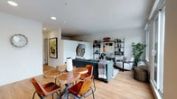 This single-floor residence showcases a thoughtfully designed open-concept layout that seamlessly blends the dining and living areas into one cohesive, inviting space. Light hardwood flooring runs throughout, lending warmth and continuity to the interior.

The dining area features a distinctive live-edge wood table paired with cognac leather chairs, bringing a natural, organic element to the space. A bold piece of framed contemporary artwork adorns the wall nearby, adding a vibrant artistic focal point.

The living area is anchored by a deep charcoal sofa and complemented by a light grey accent chair, creating a comfortable and stylish seating arrangement. A large decorative mirror and an industrial-style black shelving unit serve as standout design elements, the latter displaying books, sculptures, and curated décor that reflect a modern sensibility. A lush indoor plant in the corner adds a refreshing natural touch.

Recessed ceiling lighting provides clean, even illumination throughout, enhancing the airy feel of the space. The white walls and clean architectural lines create a neutral backdrop that allows the carefully selected furnishings and artwork to take center stage. Overall, the space exudes a contemporary yet welcoming ambiance with a well-balanced mix of industrial, natural, and modern design elements.