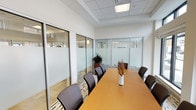 This professional conference room is thoughtfully designed to facilitate productive meetings in a polished, contemporary setting. The space is defined by its striking floor-to-ceiling glass partition walls with frosted privacy panels framed in sleek aluminum, which simultaneously provide visual openness to the surrounding office environment while maintaining a sense of enclosure and focus within the room itself.

At the heart of the space sits a generously sized rectangular conference table with a warm wood-tone finish, comfortably surrounded by cushioned dark executive chairs. A tasteful floral arrangement serves as a simple centerpiece, adding a touch of warmth to the professional atmosphere.

Natural light flows in abundantly through a row of large exterior windows fitted with adjustable roller shades, allowing for precise light control during presentations or meetings. The suspended tile ceiling features recessed flush lighting panels that provide even, bright illumination throughout.

A dedicated credenza area along one wall offers a beverage service station complete with mugs and serving trays, ideal for hosting client meetings or team gatherings. A vibrant abstract artwork displayed above adds a bold pop of color and creative energy to the otherwise neutral palette of whites and warm wood tones. The patterned area rug grounds the seating area with subtle texture, completing a refined yet welcoming professional environment.