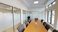 This professional conference room is thoughtfully designed to facilitate productive meetings in a polished, contemporary setting. The space is defined by its striking floor-to-ceiling glass partition walls with frosted privacy panels framed in sleek aluminum, which simultaneously provide visual openness to the surrounding office environment while maintaining a sense of enclosure and focus within the room itself.

At the heart of the space sits a generously sized rectangular conference table with a warm wood-tone finish, comfortably surrounded by cushioned dark executive chairs. A tasteful floral arrangement serves as a simple centerpiece, adding a touch of warmth to the professional atmosphere.

Natural light flows in abundantly through a row of large exterior windows fitted with adjustable roller shades, allowing for precise light control during presentations or meetings. The suspended tile ceiling features recessed flush lighting panels that provide even, bright illumination throughout.

A dedicated credenza area along one wall offers a beverage service station complete with mugs and serving trays, ideal for hosting client meetings or team gatherings. A vibrant abstract artwork displayed above adds a bold pop of color and creative energy to the otherwise neutral palette of whites and warm wood tones. The patterned area rug grounds the seating area with subtle texture, completing a refined yet welcoming professional environment.