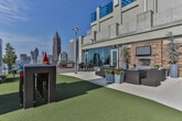 Located at 130 Sixth St NE in Atlanta's vibrant Old Fourth Ward neighborhood, this rooftop amenity deck offers an exceptional outdoor experience elevated above the city. The space is defined by expansive artificial turf that creates a lush, park-like atmosphere high above street level, providing a striking contrast to the urban skyline backdrop.

A standout feature is the stacked-stone outdoor fireplace, complete with a mounted flat-screen television and flanked by tall potted trees, creating an inviting lounge area furnished with wicker sectional seating and a large planter filled with colorful flowers. This gathering spot is ideal for relaxed socializing in any season.

The deck also showcases a dedicated seating zone with vibrant teal Adirondack chairs and a central fire pit, perfectly positioned to take in sweeping panoramic views of the Atlanta skyline, including the iconic Bank of America Plaza tower. A wicker bar-height dining table with red-cushioned stools provides additional seating for outdoor meals, while a built-in grill station and a separate dining set with dark rattan chairs offer space for full outdoor cooking and entertaining. The combination of lush greenery, thoughtfully arranged seating zones, and unobstructed city views makes this rooftop deck a dynamic and well-appointed outdoor amenity.