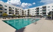 This contemporary apartment complex in Phoenix, Arizona, boasts a range of upscale amenities designed for comfortable urban living. The exterior features a clean, modern architectural style with white facades and private balconies for each unit. The property is secured by gated access, ensuring resident privacy and safety.

At the heart of the complex is a resort-style pool area, complete with a spacious deck, lounge chairs, and umbrellas for shade. Adjacent to the main pool is a smaller spa or hot tub, perfect for relaxation. The outdoor spaces are meticulously landscaped, featuring palm trees and desert-friendly plants that complement the Arizona climate.

Residents can enjoy various outdoor lounging areas, including a green space with artificial turf and comfortable seating. There's also a covered outdoor kitchen or barbecue area, ideal for social gatherings and al fresco dining. The complex offers additional recreational amenities such as a fitness center and community spaces.

The apartment buildings are multi-story structures with ample windows to allow natural light. Each unit appears to have a private balcony, providing outdoor living space and views of the surrounding area. The overall design emphasizes a blend of comfort, style, and functionality, catering to modern urban dwellers seeking a balance of luxury and convenience in their living environment.