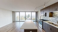 Located at 1100 Pacific Marina in Alameda, California, this contemporary apartment offers a thoughtfully designed open-concept layout bathed in natural light. The living and dining area is anchored by expansive floor-to-ceiling sliding glass doors that open to a private balcony, providing sweeping views of the surrounding treetops and marina skyline. Light-toned hardwood-style flooring runs throughout the main living spaces, complementing the clean, modern aesthetic.

The kitchen is a standout feature, equipped with full stainless steel appliances including a gas range, built-in microwave, dishwasher, and a large French door refrigerator. Rich dark-wood cabinetry extends to the ceiling, offering generous storage, while a mosaic tile backsplash adds a stylish accent. A large center island with a quartz countertop provides additional workspace and doubles as a casual dining surface. Track lighting overhead ensures the kitchen is well-lit throughout.

The bedroom is generously proportioned with the same warm flooring and crisp white walls, creating a versatile and airy retreat. The bathroom features a dark-wood vanity with a white countertop, tile flooring, and clean, functional fixtures. Overall, the unit presents a cohesive modern design with high-quality finishes throughout.