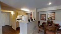 This single-story residence showcases a thoughtfully designed open-concept layout where the kitchen and dining area connect seamlessly to create a cohesive, functional living space. The kitchen is outfitted with warm honey-toned wood cabinetry that provides ample storage, complemented by a white electric range and a full-size refrigerator. A built-in range hood overhead adds a practical element above the cooking area. The light-colored countertops extend into a peninsula-style breakfast bar, which serves as a natural dividing element between the kitchen and the adjacent dining space while maintaining the open feel of the floor plan. The flooring transitions from rich, dark hardwood in the kitchen to plush carpeting in the dining area, subtly defining each zone. The dining area comfortably accommodates a full table and chair set, and decorative wall art adds a splash of color and character to the neutral-toned walls. Recessed overhead lighting in the kitchen ensures a bright, well-lit cooking environment. The overall ambiance is warm and inviting, with a practical layout designed to support everyday living and casual entertaining.