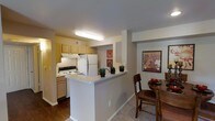 This single-story residence showcases a thoughtfully designed open-concept layout where the kitchen and dining area connect seamlessly to create a cohesive, functional living space. The kitchen is outfitted with warm honey-toned wood cabinetry that provides ample storage, complemented by a white electric range and a full-size refrigerator. A built-in range hood overhead adds a practical element above the cooking area. The light-colored countertops extend into a peninsula-style breakfast bar, which serves as a natural dividing element between the kitchen and the adjacent dining space while maintaining the open feel of the floor plan. The flooring transitions from rich, dark hardwood in the kitchen to plush carpeting in the dining area, subtly defining each zone. The dining area comfortably accommodates a full table and chair set, and decorative wall art adds a splash of color and character to the neutral-toned walls. Recessed overhead lighting in the kitchen ensures a bright, well-lit cooking environment. The overall ambiance is warm and inviting, with a practical layout designed to support everyday living and casual entertaining.