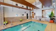 This indoor lap pool facility offers a sophisticated and well-appointed aquatic environment designed for focused swimming and fitness. The pool itself is a defined lap lane configuration, filled with clear blue-green water and marked with lane lines, catering to swimmers seeking a structured workout experience. The space is architecturally striking, featuring soaring double-height ceilings with bold, wide wood-paneled beams that add warmth and visual drama to the interior. Large glass panels at the upper level allow natural light to filter into the space, enhancing the open, airy ambiance. The pool surround is finished in neutral stone-toned tiles, providing a clean and non-slip deck area. Poolside seating, including modern white lounge chairs and a bright accent stool, offers convenient spots for rest or observation. Safety equipment, including a life ring, is prominently displayed on the wall. Tasteful decorative touches, such as a vibrant piece of wall art and a tropical potted plant, add a lively, welcoming character to the space. Pendant lighting supplements the natural light, ensuring the environment is well-illuminated at all times. A connected doorway provides access to adjacent amenity areas, making this lap pool a well-integrated part of a larger facility.