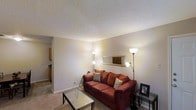 This single-floor space centers around a welcoming living room finished with wall-to-wall carpeting and neutral-toned walls that create a warm, understated atmosphere. The room features a textured popcorn ceiling that adds subtle visual depth overhead. Ambient lighting is provided by two floor lamps positioned on either side of the seating area, casting a soft, inviting glow throughout the space. A gold-framed rectangular mirror mounted on the wall serves as a decorative focal point, enhancing the sense of openness in the room. The living area comfortably accommodates a full-sized sofa and a glass-top coffee table, suggesting a practical yet comfortable layout suited for everyday living. A small side table near the sofa provides additional surface space. A partial view into an adjacent kitchen area is visible from the living room, revealing wooden cabinetry and a connected layout that supports easy flow between spaces. The overall ambiance of the space is cozy and functional, with warm lighting and simple finishes contributing to an approachable and livable environment.