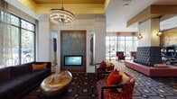 Located at 3833 S Bond Ave in Portland, Oregon, this thoughtfully designed lobby space makes a striking first impression with its bold, eclectic interior aesthetic. The single-floor layout is anchored by a dramatic mosaic-tiled fireplace framed in rich wood, serving as a focal point for one of several distinct seating areas throughout the space. Overhead, tiered crystal chandeliers and pendant lighting cast a warm glow that complements the gold-accented ceiling details and textured wall treatments.

The main lounge area is furnished with vibrant patterned armchairs in red and black floral upholstery, a sculptural bronze coffee table, and a dark velvet sofa positioned to take full advantage of the floor-to-ceiling windows. These expansive windows flood the space with natural light while offering views of the surrounding streetscape. Sheer drapes add a soft, layered touch to the glazed facades.

A secondary lounge features a tall tufted charcoal banquette backed by a gilded mosaic wall panel, with a large flat-screen television nearby for entertainment. A dedicated business center with a workstation and ergonomic seating provides a functional retreat. A bold red-accented island seating piece serves as a central divider, adding a pop of color to the open floor plan while defining separate zones within the cohesive, design-forward environment.