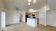 Located at 7702 Little Fox Ln in Wellington, Colorado, this single-floor residence offers a well-designed, open-concept layout that seamlessly connects the living and kitchen areas. The kitchen is a functional centerpiece, featuring light wood cabinetry that extends to the ceiling for generous storage, a dark contrasting countertop, and black appliances including a refrigerator with ice dispenser, a built-in microwave, and a dishwasher. A peninsula island with a raised bar provides a natural division between the kitchen and the living space, making it ideal for casual dining or entertaining. The living area is finished with neutral wall-to-wall carpeting that adds warmth underfoot, while the kitchen transitions to tile flooring for easy maintenance. Ceiling fans are present in the living area, ensuring year-round comfort. Soft, ambient lighting from a chandelier-style fixture and under-cabinet lighting enhances the overall atmosphere. A hallway visible from the main living area leads to additional rooms, and the layout includes interior doors with classic paneled detailing, contributing to a clean and cohesive aesthetic throughout the home.
