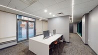 This contemporary single-floor amenity space is thoughtfully designed to blend functionality with leisure. The entry area features a large, clean-lined white workstation desk equipped with computer monitors and seating for multiple users, positioned near floor-to-ceiling glass entry doors that allow natural light to filter in from the street. Recessed lighting and sleek linear LED strips run along the drop tile ceiling throughout, providing consistent, modern illumination across the space.

A striking textured stone accent wall serves as a bold architectural focal point in the transitional area, adding warmth and visual depth against the otherwise neutral grey palette. Adjacent to it, a lush potted plant introduces a touch of greenery.

The recreation lounge area is a standout, anchored by a stylish pool table beneath a pair of large matte pendant lights. A spacious grey sectional sofa faces two deep teal-upholstered wall niches, creating a dramatic and inviting backdrop. Warm orange accent chairs add a pop of color in a separate seating nook.

A dedicated lounge area features a large-screen TV mounted above a long, modern linear fireplace framed in dark stone, with a plush sofa and patterned area rug completing the relaxed atmosphere. Roller shades along the windowed wall provide privacy and light control throughout.