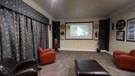 This dedicated home movie theater is thoughtfully designed to deliver an authentic cinematic experience. The room is anchored by a large wall-mounted projection screen that serves as the focal point, ideal for movie nights and entertainment gatherings. Crown molding runs along the perimeter of the ceiling, adding a refined architectural touch that elevates the overall aesthetic of the space.

The walls are adorned with bold, patterned floor-to-ceiling drapes in a deep, dark tone that effectively block ambient light and enhance the theatrical atmosphere. Decorative film reel wall accents flank the projection screen, reinforcing the cinema theme with a charming, nostalgic nod to classic moviemaking.

Seating includes plush leather armchairs and an ottoman, offering comfortable viewing positions throughout the room. The carpeted flooring adds warmth underfoot and contributes to the room's excellent acoustics. In-ceiling speakers are integrated seamlessly into the space, supporting a full surround sound experience. A classic red popcorn machine adds a fun, authentic touch that rounds out the theater ambiance. Wall-mounted sconces provide subtle accent lighting, allowing for a perfectly dimmed environment during screenings. Overall, this space is a well-appointed, immersive home theater designed for both comfort and style.
