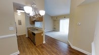 This single-floor residence showcases an open-concept layout that seamlessly connects the kitchen and living areas, creating a cohesive and functional living environment. The kitchen is appointed with warm-toned wood cabinetry, a stainless steel dishwasher, and a countertop sink, all illuminated by an elegant multi-bulb chandelier that serves as a central design feature. The flooring transitions from rich hardwood in the kitchen and dining area to plush carpeting in the living space, adding a sense of comfort and distinction between zones. The living area benefits from generous natural light streaming through a full-length sliding glass door dressed with vertical blinds, which also provides access to an outdoor area. Vaulted ceilings contribute to an airy, spacious ambiance throughout the unit, enhancing the overall sense of openness. Neutral beige walls provide a versatile backdrop that complements the warm wood tones and flooring throughout. HVAC vents are thoughtfully placed for efficient climate control. The layout is well-suited for everyday living, offering a practical flow between cooking, dining, and relaxing spaces within a bright and welcoming interior.