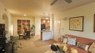 This single-floor residence offers an open-concept design that effortlessly connects the living, dining, and kitchen areas into one cohesive and functional space. The living area features warm neutral tones, plush carpeting, and a ceiling fan overhead, creating a comfortable and inviting atmosphere. A cozy sofa with decorative throw pillows anchors the space, complemented by tasteful wall art that adds a personal touch.

The kitchen is positioned centrally and defined by a breakfast bar counter that doubles as a natural divider between living and cooking spaces. Warm wood cabinetry, pendant lighting, and a black refrigerator give the kitchen a classic and practical character. Tile flooring in the kitchen area contrasts nicely with the soft carpeting of the surrounding rooms.

Adjacent to the kitchen, the dining area accommodates a full dining table and chairs, bathed in warm ambient light. Lush indoor plants throughout the space bring a refreshing natural element to the interior. The entryway, visible from the main living area, features a solid front door with a security panel, ensuring both convenience and peace of mind. Overall, the space exudes a warm, lived-in charm with a thoughtful layout designed for comfortable everyday living.