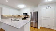 This thoughtfully designed 1-bedroom, 1-bathroom residence offers a clean, contemporary aesthetic across a single floor. The open-concept kitchen and living area serves as the heart of the home, showcasing white shaker-style cabinetry, granite countertops with subtle grey veining, a tiled backsplash, and a full suite of stainless steel appliances including a refrigerator, electric range, over-the-range microwave, and dishwasher. A peninsula with a built-in sink provides additional prep space and creates a natural division between the kitchen and the living area. Warm hardwood flooring flows seamlessly throughout the main living spaces, complementing the bright, neutral palette of the walls and trim. The bedroom is generously sized, offering ample room for furnishings, and features a large window that brings in natural light and views of the surrounding neighborhood. It connects directly to a dedicated closet with built-in shelving for organized storage. The bathroom is finished with large-format white tiles and a soaking tub with a decorative vertical mosaic tile accent strip and chrome fixtures. A dedicated entrance and hallway complete the functional layout of this well-organized residence.