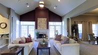 This single-story home showcases a striking living room as its centerpiece, distinguished by soaring vaulted ceilings that create an expansive, airy atmosphere. A bold deep plum accent wall draws the eye upward, emphasizing the dramatic ceiling height and serving as a commanding backdrop for the white-mantled fireplace below. Flanking the fireplace, large white-framed windows allow generous natural light to fill the space, enhancing the room's open and welcoming character. The living area is furnished with two cream-colored sofas arranged symmetrically around a dark wood coffee table, anchored by a patterned area rug that adds texture and visual interest. Decorative throw pillows in complementary burgundy and purple tones tie the color palette together cohesively. Recessed lighting, along with a ceiling fan and central light fixture, provides layered illumination throughout. Decorative elements including potted plants, lanterns, and artwork contribute warmth and personality. Architectural columns visible toward the rear of the space hint at additional rooms beyond, suggesting a thoughtfully designed, connected floor plan throughout this single-level home.
