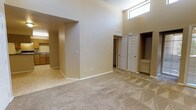 This single-floor space is characterized by its notably high ceilings, which lend an airy, open feel to the interior. A clerestory window positioned near the top of the wall allows natural light to filter into the main living area, enhancing the sense of height and brightness throughout the space.

The main living area features wall-to-wall carpeting in a neutral beige tone, creating a warm and comfortable atmosphere. The flooring transitions to tile in the adjoining kitchen area, which is visible through a wide, open passageway. The kitchen includes wooden cabinetry with a warm finish, a sink, and sufficient counter space, offering a functional layout.

Neutral-toned walls throughout the space contribute to a clean and cohesive aesthetic. Two white paneled doors are visible from the main living area, suggesting access to additional rooms such as bedrooms or bathrooms. A recessed niche near one of the doorways provides a built-in storage or display area, adding a practical architectural detail.

The open flow between the living and kitchen areas, combined with the generous ceiling height and natural light, gives the space a welcoming and versatile character suitable for a variety of uses.